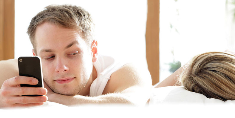 Homem na cama com o celular e olhando para a mulher dormindo